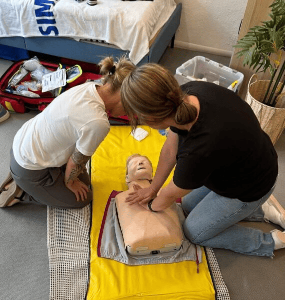 Zwei Frauen knien auf einer gelben Matte und führen eine Herzdruckmassage an einer HLW-Übungspuppe durch.