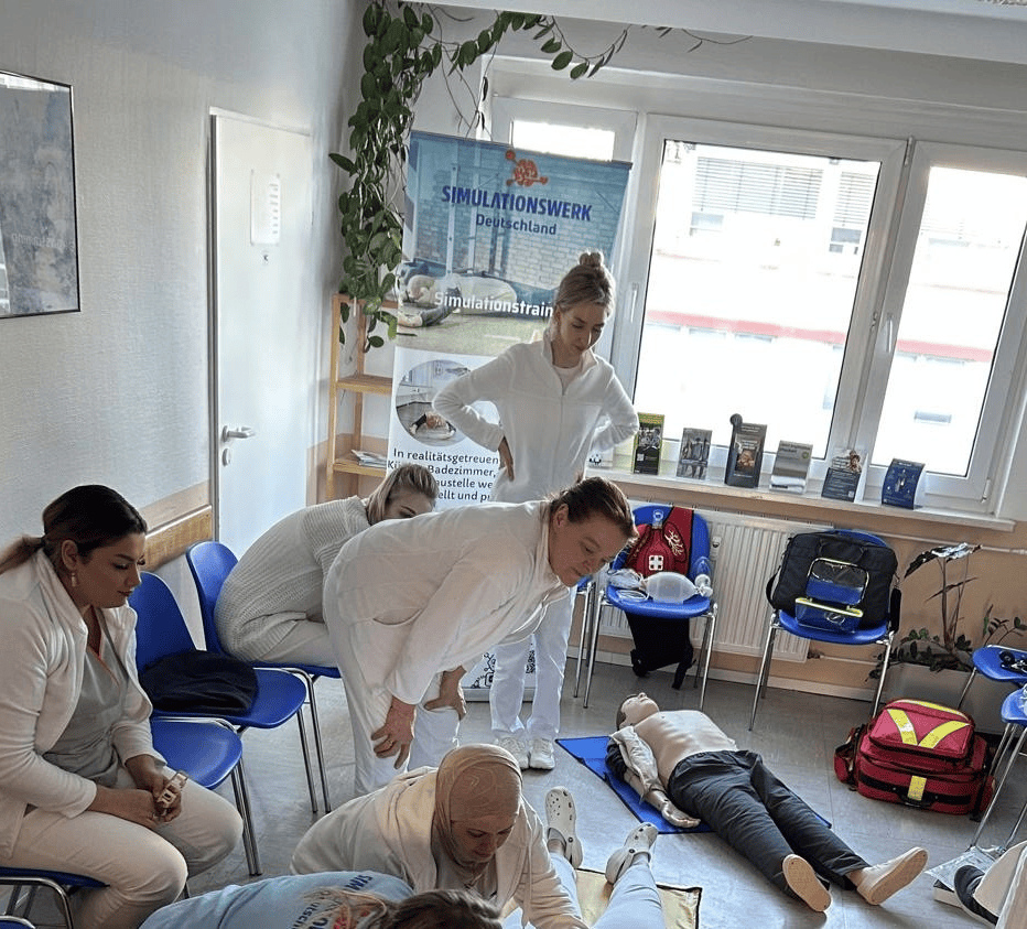 Eine Gruppe von Teilnehmern Arztpraxis in weißer Berufsbekleidung während einer Erste-Hilfe-Simulation Notfalltrainig mit einer HLW-Puppe in einem hellen Schulungsraum.